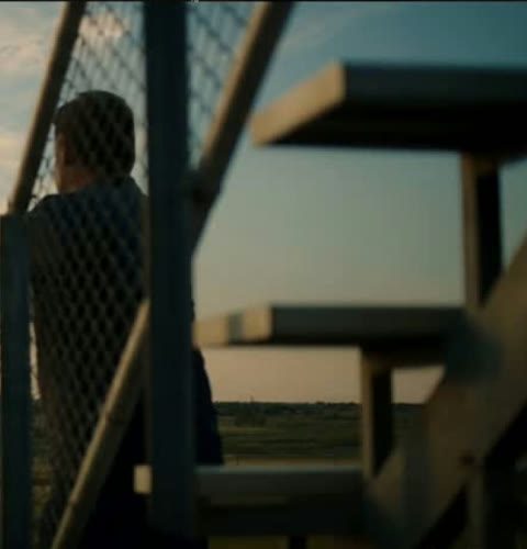 The Higgsfield generation. The actor stands on the bleachers behind a chain-link fence, sunset light across the track behind him, camera move matched to the original handheld.